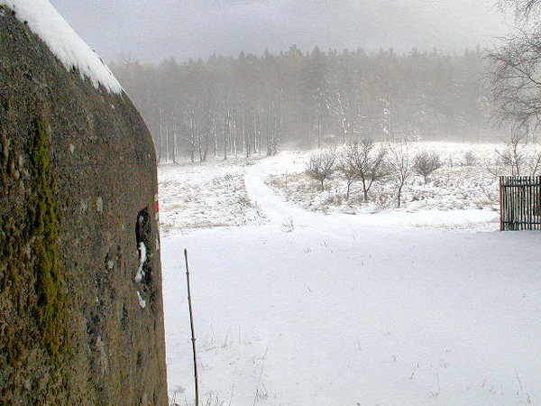Pohled od řopíku L1/52/A160 v Horním Sedle přes louku k sousednímu objektu č.53, prosvítajícímu mezi stromy.