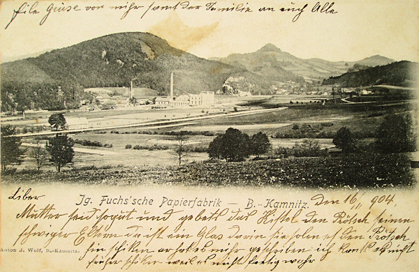 Diese Ansichtskarte zeigt die Papierfabrik im Tal oberhalb von Horní Kamenice (Oberkamnitz). Hinter dem Fabriksgebäude ist der Břidličný vrch (Schiefereberg) und rechts von ihm der spitzige Střední vrch (Mittenberg).