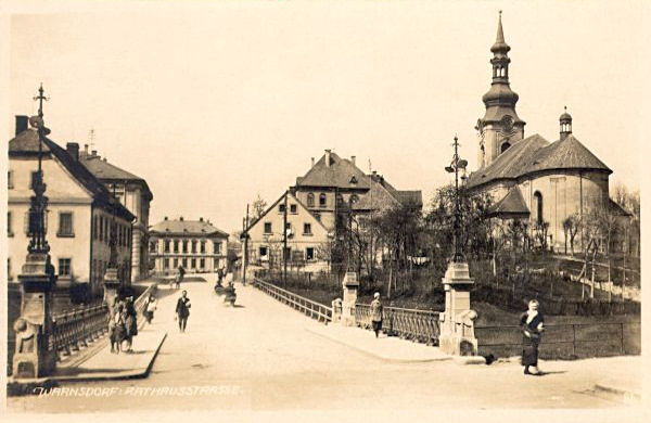 Auf dieser Ansichtskarte aus den Jahren zwischen den beiden Weltkriegen sieht man die Rathausbrücke (heute: Most Legií) von 1902 und einen Teil der zum Stadplatz führenden Rathausstrasse. Rechts ist die Kirche St. Petri u. Pauli.