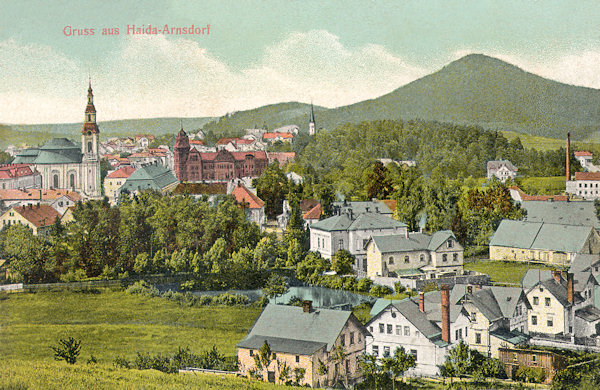 This picture postcard from the first years of the 20th century shows the town centre with the church Assumption of the Virgin and the monumental building of the former savings bank. The houses in the foreground belong to the village Arnultovice which gradually merged with Nový Bor. In the background to the left there is the Chotovický vrch-hill.