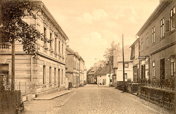 Na této pohlednici vidíme dolní část Rumburské ulice (nynější Komenského), která ve 30. letech 20. století přestala vyhovovat rostoucímu provozu a byla proto podstatně rozšířena. Zbořena přitom byla většina domů na pravé straně ulice, s výjimkou nejbližších dvou.