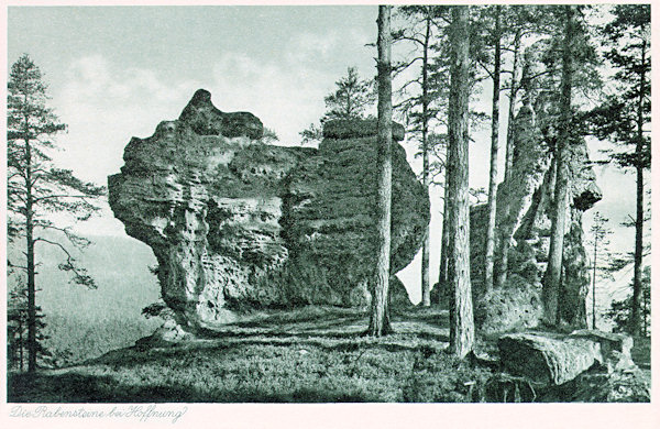 This picture postcard shows a nonstandard wiew of the sandstone rocks over the southern border of the village Naděje. Whereas the smaller rock in the foregrund we see distinctly, the more famous rock Křížová věž which is the subject of numerous folk tales is prevailingly hidden behind the trees.