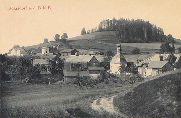 Tato pohlednice zachycuje střední část obce s kaplí Nejsvětější Trojice, postavenou v roce 1788. Za domky vlevo je nádraží a nad ním vrch Sokolík.