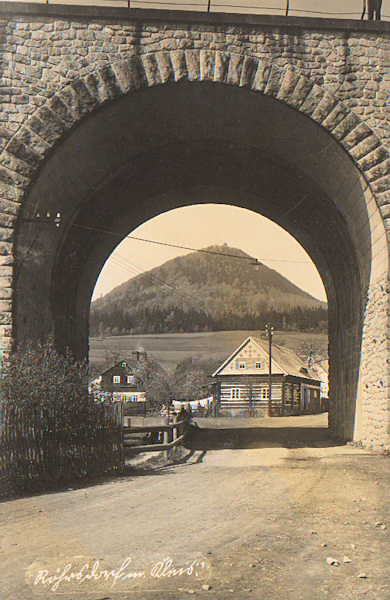 Tato pohlednice zachycuje pod portálem železničního mostu několik domků v horní části obce a horu Klíč v pozadí.